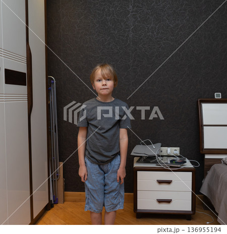 A young blonde boy stands straight facing the camera in a bedroom setting. He is wearing a grey t-shirt and blue denim shorts. A young blonde boy stands straight facing the camera in a bedroom setting. He is wearing a grey t-shirt and blue denim shorts. 136955194