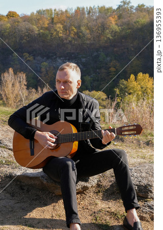 Portrait of smiling caucasian artist plays wooden acoustic guitar in autumnal park. Young songwriter plays string musical instrument hobby outside in nature fall time. Audio music healing Portrait of smiling caucasian artist plays wooden acoustic guitar in autumnal park. Young songwriter plays string musical instrument hobby outside in nature fall time. Audio music healing 136955393