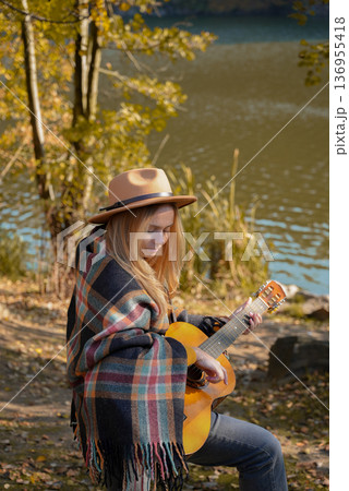 Portrait of smiling caucasian artist plays wooden acoustic guitar in autumnal park. Young songwriter plays string musical instrument hobby outside in nature fall time. Audio music healing Portrait of smiling caucasian artist plays wooden acoustic guitar in autumnal park. Young songwriter plays string musical instrument hobby outside in nature fall time. Audio music healing 136955418