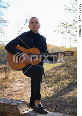Portrait of smiling caucasian artist plays wooden acoustic guitar in autumnal park. Young songwriter plays string musical instrument hobby outside in nature fall time. Audio music healing Portrait of smiling caucasian artist plays wooden acoustic guitar in autumnal park. Young songwriter plays string musical instrument hobby outside in nature fall time. Audio music healing 136955435