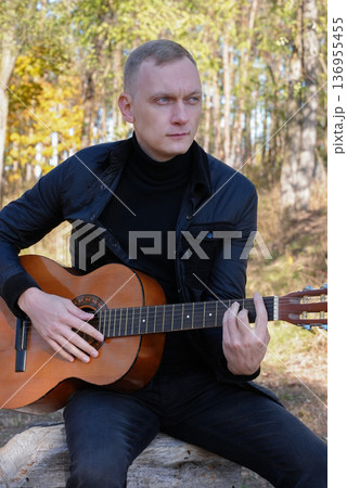 Beautiful blonde man playing acoustic guitar and singing relaxing enjoying outdoors on sunny autumn day. Artist learning and practicing playing musical instrument. Fall leaves autumnal 136955455