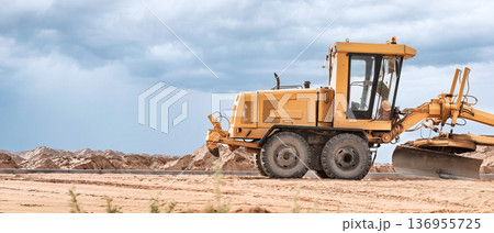 A yellow grader is actively leveling the sandy terrain at a construction site while dramatic clouds loom in the sky during late afternoon 136955725