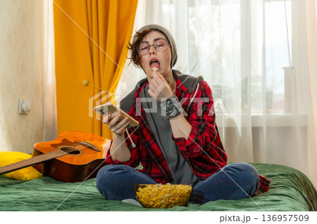 Caucasian teenage student woman watching television at home. She relaxes on bed with popcorn. Entertainment concept. Mid shot Caucasian teenage student woman watching television at home. She relaxes on bed with popcorn. Entertainment concept. Mid shot 136957509