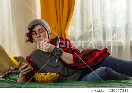Caucasian teenage woman student resting indoors during free time. Girl eats popcorn and using smartphone. Home comfort theme. Mid shot Caucasian teenage woman student resting indoors during free time. Girl eats popcorn and using smartphone. Home comfort theme. Mid shot 136957511