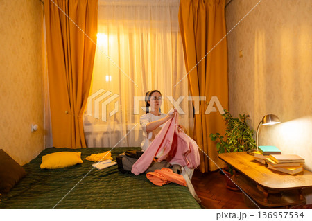 Caucasian woman folding clothes indoors during evening light. She keeps her room tidy. Clean living space theme. Mid shot Caucasian woman folding clothes indoors during evening light. She keeps her room tidy. Clean living space theme. Mid shot 136957534