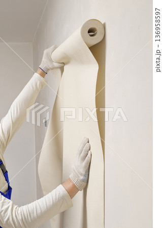 Vertical view a woker holds a roll of vinyl wallpaper against the wall. 136958597