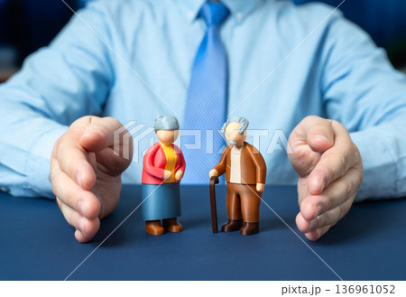 A man protects with his hands the figurines of a couple of elderly pensioners. Social protection and financial security. Support for seniors and pension insurance policy A man protects with his hands the figurines of a couple of elderly pensioners. Social protection and financial security. Support for seniors and pension insurance policy 136961052