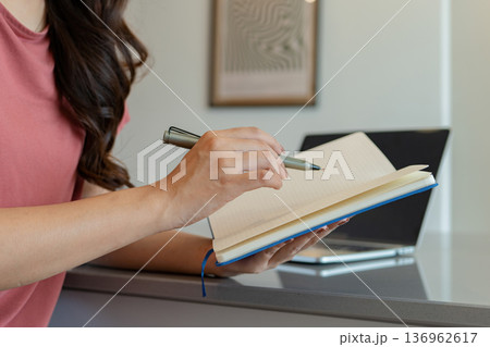 Smiling Asian Woman Writing in Notebook at Kitchen Counter 136962617