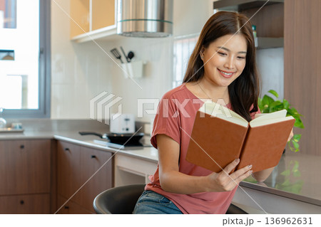 Smiling Asian Woman Reading Book with Coffee in Kitchen 136962631