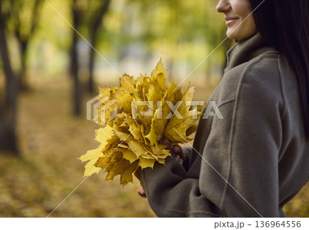 Close-up of young woman in coat holding autumn leaves bouquet in city park Close-up of young woman in coat holding autumn leaves bouquet in city park 136964556