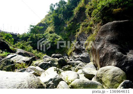 Sunlit rocky riverbed with large boulders and lush green mountain slopes 136964631