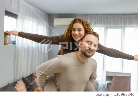 Happy couple having fun at home man giving woman piggyback ride in living room 136965346