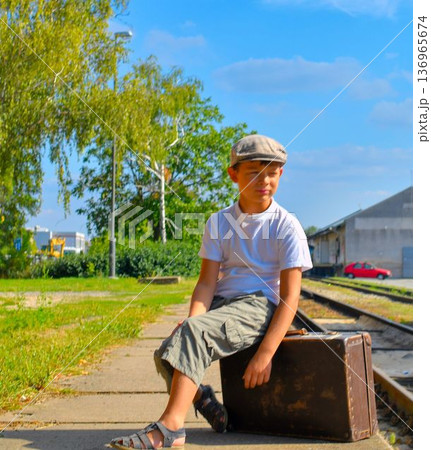 A cute little boy sits on an old suitcase. The adorable boy in a flat cap waits on the station platform for the holiday train. The concept of a carefree childhood, travel, adventure, summer holidays 136965674
