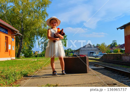 A cute little girl in a white dress with a stuffed toy and an aged luggage. The adorable smiling girl is standing on a train station platform. The concept of the end of summer holidays, travel, and A cute little girl in a white dress with a stuffed toy and an aged luggage. The adorable smiling girl is standing on a train station platform. The concept of the end of summer holidays, travel, and 136965737