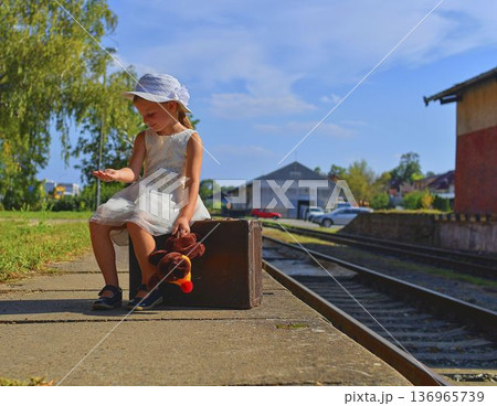 A cute girl at the train station. A little girl holds a plush toy and sits on an old suitcase. The concept of travel, vacations, and a carefree childhood. Retro and vintage vibes. A cute girl at the train station. A little girl holds a plush toy and sits on an old suitcase. The concept of travel, vacations, and a carefree childhood. Retro and vintage vibes. 136965739