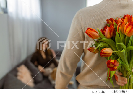 Man hiding bouquet of tulips behind back preparing romantic surprise at home Man hiding bouquet of tulips behind back preparing romantic surprise at home 136969672