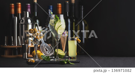Bar composition featuring a variety of cocktails: martini with olives, citrus mint highball, and limoncello shot against wine bottles, shaker, strainer and fresh ingredients Bar composition featuring a variety of cocktails: martini with olives, citrus mint highball, and limoncello shot against wine bottles, shaker, strainer and fresh ingredients 136971082
