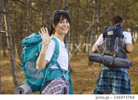 Smiling female hiker with backpack waving while partner walking on forest trail 136973768