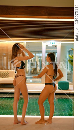 Two women organize shots amidst tropical plants at hotel poolside 136975154