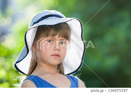Close-up portrait of serious little girl in a big hat. Child having fun time outdoors in summer. 136975597