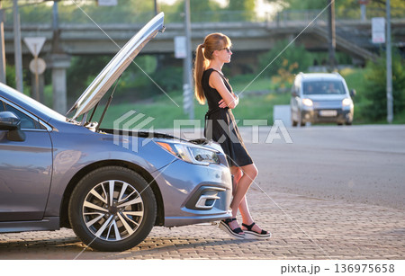 Helpless woman standing near her car with open bonnet inspecting broken motor. Young female driver having trouble with vehicle Helpless woman standing near her car with open bonnet inspecting broken motor. Young female driver having trouble with vehicle 136975658