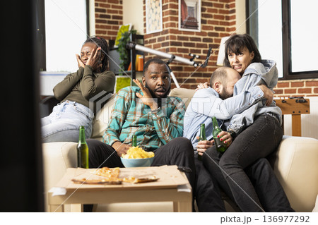 Frightened friends watch horror movie together in apartment, reacting to shocking scene with fear and nervous expressions. Caucasian couple hugs as diverse group bonds over thriller film at home. 136977292