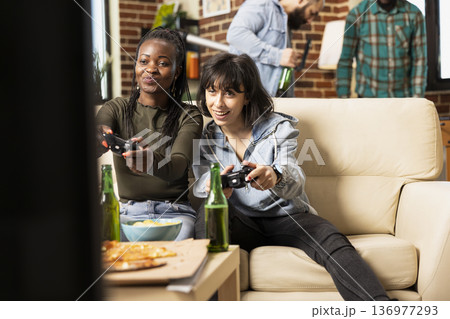 Young women with wireless controllers, cheerfully playing video games and enjoying leisure time at home. Weekend relaxation in cozy apartment as diverse female friends participate in online gaming. 136977293