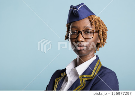 Professional cabin crew member looks at camera, posing confidently in studio. Portrait of elegant black woman in aviation uniform and glasses, representing female confidence and leadership. Professional cabin crew member looks at camera, posing confidently in studio. Portrait of elegant black woman in aviation uniform and glasses, representing female confidence and leadership. 136977297