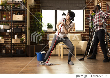 White woman with headphones sings and dances in living room, turning household chores into fun performance. Diverse couple enjoys apartment cleanup as wife playfully imitates rockstar with mop in hand White woman with headphones sings and dances in living room, turning household chores into fun performance. Diverse couple enjoys apartment cleanup as wife playfully imitates rockstar with mop in hand 136977306