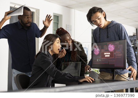 Diverse colleagues show frustration and focus while analyzing system crash on computer screen in tech startup workspace. Agile development team collaborates to solve complex programming bug. Diverse colleagues show frustration and focus while analyzing system crash on computer screen in tech startup workspace. Agile development team collaborates to solve complex programming bug. 136977390