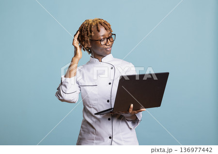 Black woman chef browses internet for fresh menu inspiration and cookbook references. African american female cook checking new meal ideas and preparing updated kitchen plans for professional service. 136977442
