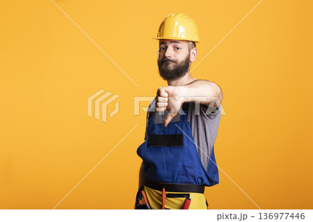 Disappointed male contractor signals disapproval with thumbs down. Man wearing protective gear stands in studio, ideal for concepts of poor results, rejection, low ratings or construction setbacks. Disappointed male contractor signals disapproval with thumbs down. Man wearing protective gear stands in studio, ideal for concepts of poor results, rejection, low ratings or construction setbacks. 136977446