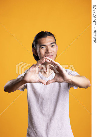 Happy gentleman makes heart symbol with fingers while smiling at camera. Friendly gesture expresses warmth, affection and kindness, representing love and positive communication in cheerful portrait. 136977479