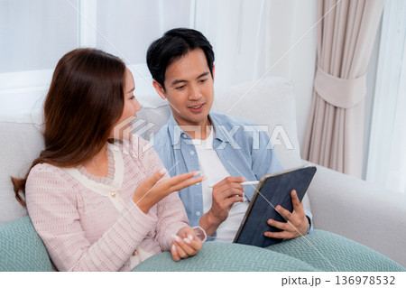 Happy asian couple planning and writing notes on digital tablet device together sitting on sofa. Happy asian couple planning and writing notes on digital tablet device together sitting on sofa. 136978532