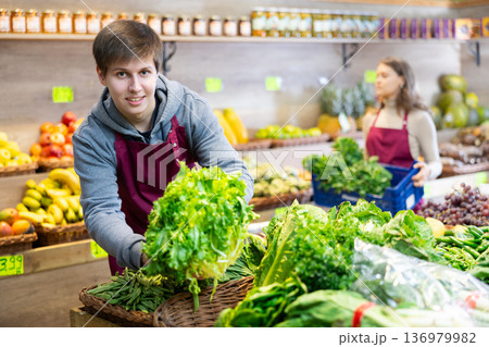 Young male seller putting lettuce in vegetable basket in grocery market Young male seller putting lettuce in vegetable basket in grocery market 136979982