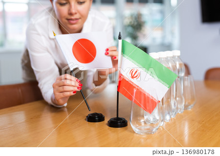 A woman in a white blouse with friendly smile arrangs flags of Japan and Iran for meeting. A woman in a white blouse with friendly smile arrangs flags of Japan and Iran for meeting. 136980178