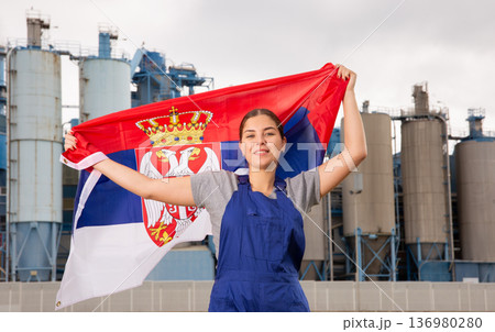 Happy young woman worker with flag of Serbia against background of factory Happy young woman worker with flag of Serbia against background of factory 136980280