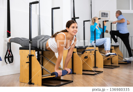 Young European woman working out on Pilates Wanda chair machine during group class with elderly people in gym Young European woman working out on Pilates Wanda chair machine during group class with elderly people in gym 136980335