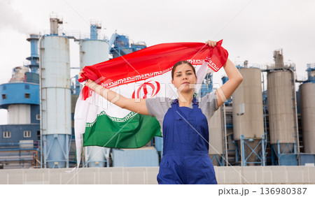 Calm young woman worker with flag of Iran against background of factory 136980387