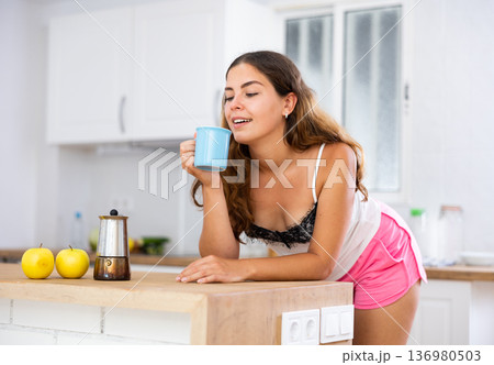 Portrait of young woman leaning on table in kitchen and drinking coffee 136980503