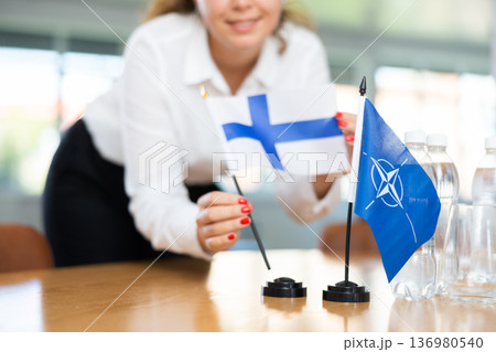 Female secretary places flags of the NATO (OTAN) and Finland on the table before negotiations of top political figures 136980540