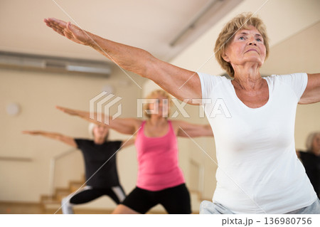 Group of aged women standing in warrior II pose in fitness room Group of aged women standing in warrior II pose in fitness room 136980756