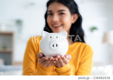 Young Asian woman holding piggy bank and sitting on couch in living room at home 136982016