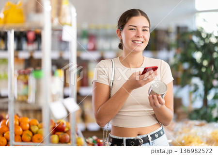 European girl customer at shop scans QR code on tin cans of preserves using phone camera 136982572