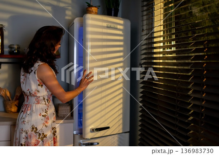 Woman seeks comfort in kitchen, Ambient scene of woman retrieving snack in dim light, Calm apartment setting with floral dressed woman exploring refrigerated items at night 136983730