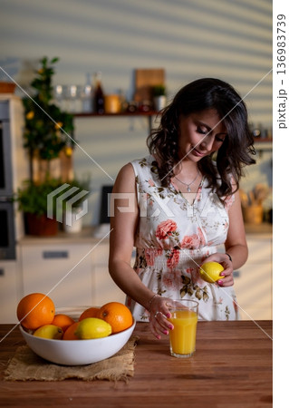 Preparing healthy breakfast drink, Woman while pouring fresh orange juice in kitchen setting, Woman in floral dress creates nutritious orange juice in cozy kitchen environment Preparing healthy breakfast drink, Woman while pouring fresh orange juice in kitchen setting, Woman in floral dress creates nutritious orange juice in cozy kitchen environment 136983739