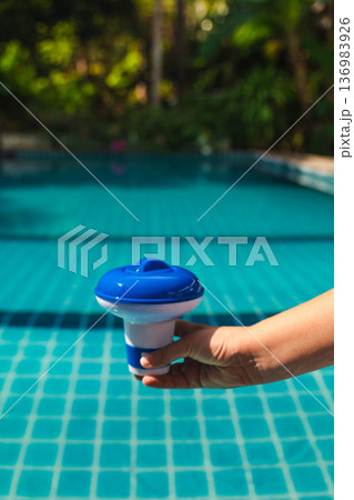 Floating chlorine dispenser held by hand above swimming pool water, device used for slow release of pool chemicals, outdoor pool maintenance 136983926