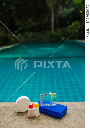 Pool water testing kit with chlorine tablet, reagent bottles and color scale on stone edge, tropical pool background 136983927