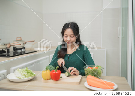 Young Woman Preparing Fresh Vegetables in Modern Kitchen, Slicing Cucumbers with Colorful Ingredients on Table, Healthy Cooking Concept 136984520