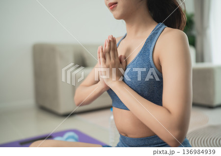 Young woman meditating indoors with folded hands while practicing yoga in sportswear, showcasing calmness and focus in a serene living space environment 136984939